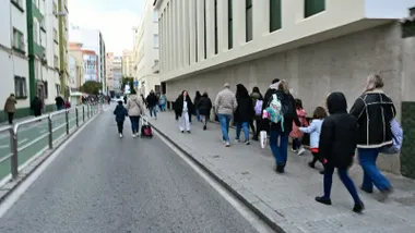 Grupo de niños y niñas caminando junto a sus padres y madres por la acera de la calle Trille, sin vehículos debido al corte temporal de tráfico. Al fondo se ve la entrada del colegio Rebaño de María, con señalización y vallas que delimitan el itinerario escolar seguro.