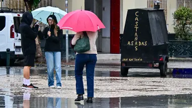 Dos jóvenes se toman una foto bajo un paraguas mientras un mujer camina por la Plaza de San Antonio de Cádiz junto al puesto de castañas asadas.