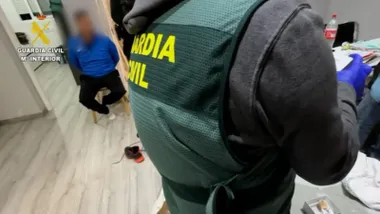 Agente de la Guardia Civil en primer término con un detenido con sudadera azul sentado y esposado en una silla.