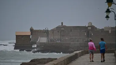 Imagen del Paseo Fernando Quiñones en Cádiz durante la borrasca Claudia: el mar aparece muy embravecido, el cielo completamente nublado y con lluvia. Dos personas, una en pantalón corto y otra en camiseta, avanzan por el paseo mientras se ve al fondo el castillo de San Sebastián.