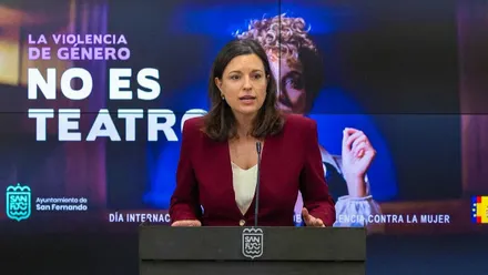 Patricia Cavada con chaqueta burdeos y una pantalla con el lema "No es teatro" referente al 25N.