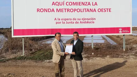 José María Román y José Vera posan junto a un cartel blanco y rosa de grandes proporciones de la Ronda Oeste de Chiclana.