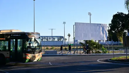 Imagen del solar vacío donde está prevista la construcción del nuevo hospital de Cádiz. En primer plano se ve un autobús circulando y varias personas caminando por la acera junto a una valla metálica que delimita el terreno.