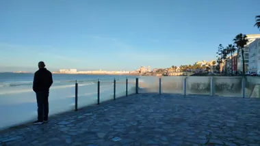 Imagen tomada desde la zona de calistenia de Santa María del Mar, en Cádiz. Se los paneles de metacrilato del mirador, mojados por la humedad de primera hora. Un hombre de pie mira hacia el casco antiguo, visible al fondo con la catedral destacando bajo un cielo ya despejado.