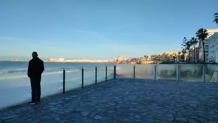 Imagen tomada desde la zona de calistenia de Santa María del Mar, en Cádiz. Se los paneles de metacrilato del mirador, mojados por la humedad de primera hora. Un hombre de pie mira hacia el casco antiguo, visible al fondo con la catedral destacando bajo un cielo ya despejado.