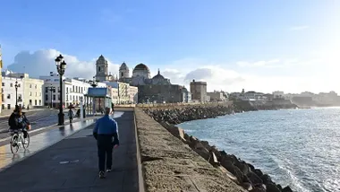 Un hombre camina y una bici circula por el carril bici del Campo del Sur de Cádiz en una mañana soleada con la catedral al fondo