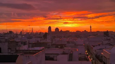 Imagen tomada al amanecer en Cádiz capital. El cielo aparece cubierto de tonos intensos naranjas y rojizos, fruto de la mezcla de humedad y nubes en proceso de disipación. En primer plano se distinguen las siluetas de la Catedral de Cádiz, el pirulí de comunicaciones y el segundo puente, recortadas sobre un cielo luminoso que anuncia una progresiva mejoría del tiempo.