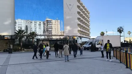 Varias personas caminan por la glorieta Ana Orantes en Cádiz, una zona peatonal amplia con árboles y tráfico alrededor, captada en un día luminoso. La escena muestra el movimiento cotidiano de la ciudad.