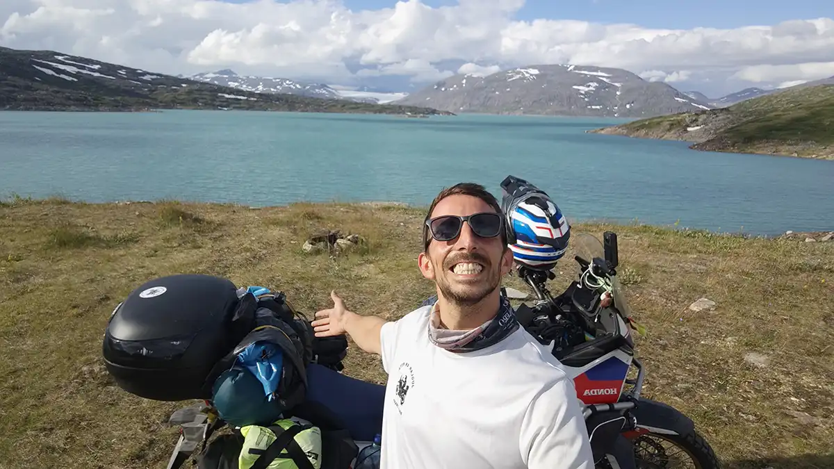 arlos Romero aparece sonriente junto a su moto frente a un lago rodeado de montañas. La imagen refleja el equilibrio entre esfuerzo, soledad y conexión con la naturaleza durante su viaje en solitario por Europa.