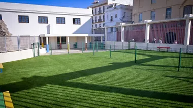Imagen de uno de los patios de la Escuela Infantil Municipal Santa María, con suelo de césped artificial verde recién instalado, varias zonas de juego infantil con elementos de colores y espacios amplios y seguros. El entorno está vallado y adaptado para niños pequeños, con acceso sencillo y sin obstáculos, pensado para el juego y la actividad al aire libre.