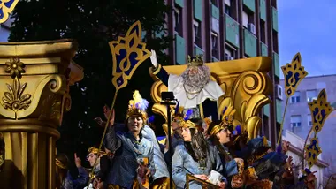 El Rey Melchor con corona y barba blanca saluda desde una carroza dorada durante la Cabalgata de Cádiz de 2026, acompañado por varios integrantes del cortejo.