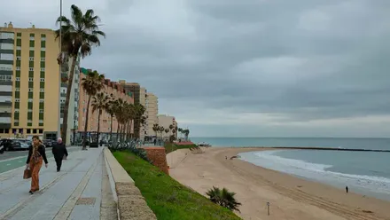 Dos personas caminan por el Paseo Marítimo de Cádiz bajo un cielo gris y con el espigón de Santa Maróia del Mar y la playa al fondo. Foto: José Luis Porquicho Prada.