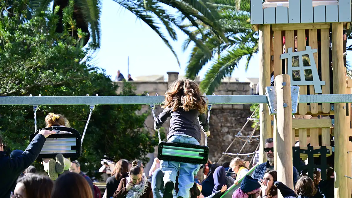 Imagen de varios niños y niñas columpiándose en un parque infantil, con expresiones de alegría y movimiento. Los columpios están ocupados y alrededor se aprecia un ambiente familiar, con otros menores jugando y adultos observando desde zonas cercanas.