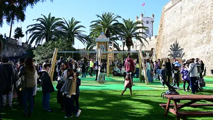 Imagen panorámica de un parque infantil lleno de niños y niñas jugando en columpios y zonas de recreo, con familias sentadas en mesas de picnic y paseando. Al fondo se distingue el Torreón de las Puertas de Tierra, integrado en el entorno urbano, bajo un cielo despejado y con un ambiente animado y alegre.