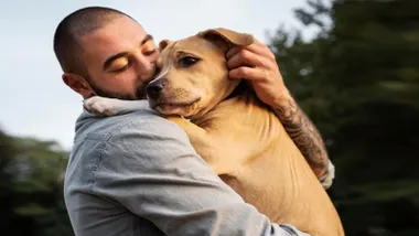 Hombre abrazando a un perro, lo tiene en brazos.