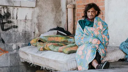 Hombre enrollado en una manta sentado en un colchón, duerme en la calle.