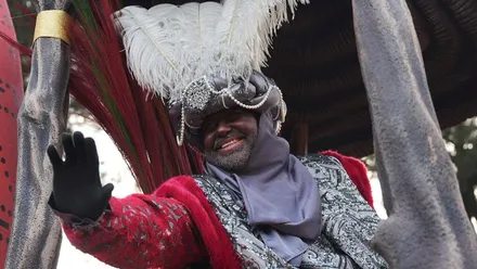 Rey negro con gorro de plumas blancas y túnica roja con fular en tonos plomo.