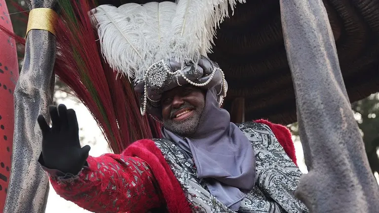 Rey negro con gorro de plumas blancas y túnica roja con fular en tonos plomo.
