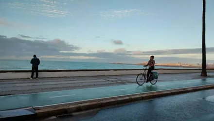 Una mujer circula en su bici por la zona de Santa María del Mar en un día que ha amanecido con suelo mojado y cielo casi despejado en Cádiz. Foto: José Luis Porquicho Prada.