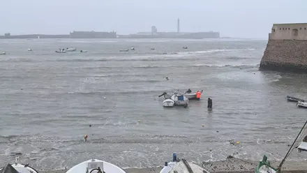 Tres personas trabajan bajo la lluvia en la playa de La Caleta para poner a salvo varias embarcaciones afectadas por el temporal. En primer plano se observa una barca medio hundida junto a otras pequeñas embarcaciones. El ambiente es húmedo y lluvioso, con bruma en el aire. Al fondo se distingue el castillo de San Sebastián y, a la derecha de la imagen, el castillo de Santa Catalina, con el mar alterado por el viento.