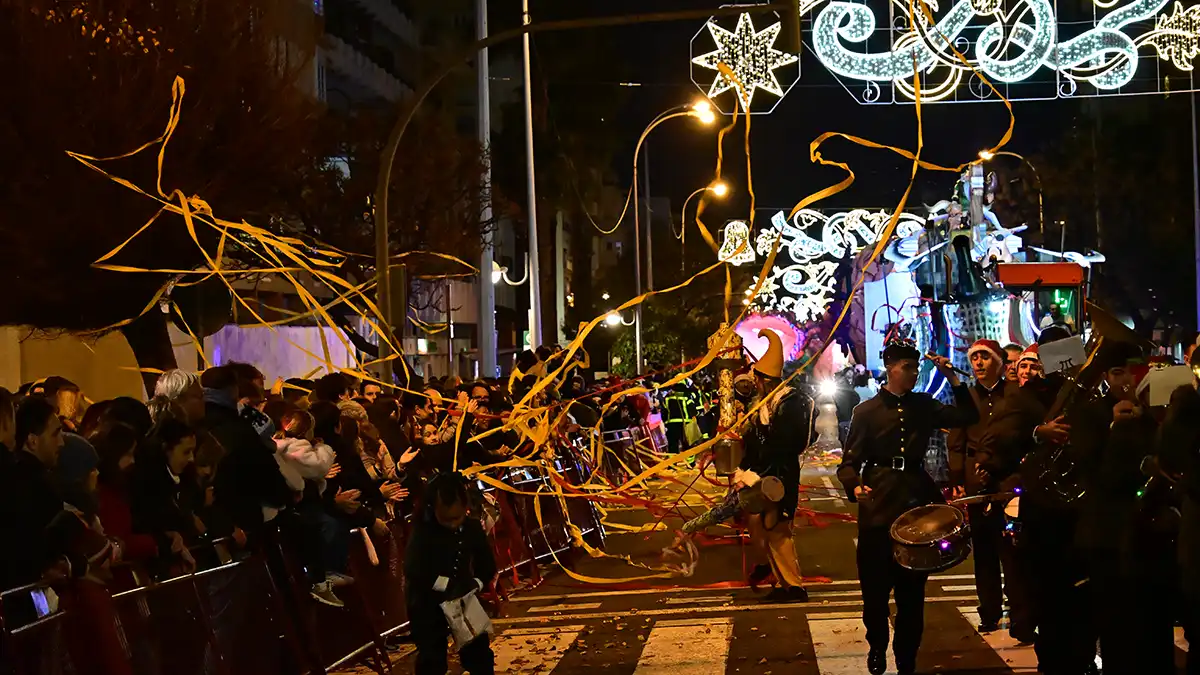 Banda de música desfilando y tocando instrumentos durante la Cabalgata de Reyes Magos, con público a ambos lados de la calle.