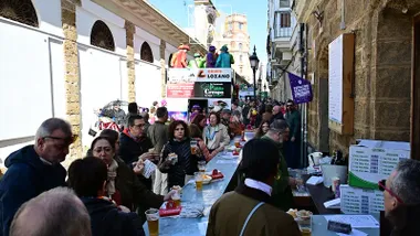 Imagen del Carnaval de Cádiz en la calle: varias personas consumen en una barra instalada al aire libre mientras, a pocos metros, un coro canta desde su batea decorada durante el carrusel, rodeado de público que disfruta del ambiente festivo.