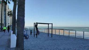 Un joven se ejercita en la zona de Calistenia en Santa María del Mar mientras una pareja camina por el Paseo Marítimo en este viernes despejado y soleado. Foto: José Luis Porquicho Prada.