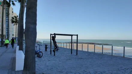 Un joven se ejercita en la zona de Calistenia en Santa María del Mar mientras una pareja camina por el Paseo Marítimo en este viernes despejado y soleado. Foto: José Luis Porquicho Prada.
