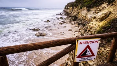 Barrera con un cartel de peligro por derrumbe en la playa de Caños de Meca en Barbate, se ve la playa en una situación de poca arena y muchas rocas.