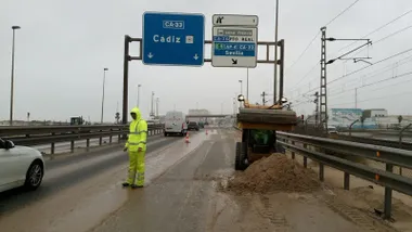 Uno de los carriles de la autobía entre Cádiz y San fernando con gran acumulación de arena mientras operarios trabajan en impiarla y los coches siguen circulando por el otro carril