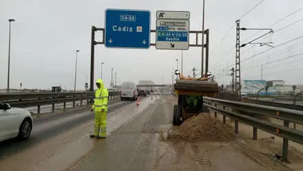 Uno de los carriles de la autobía entre Cádiz y San fernando con gran acumulación de arena mientras operarios trabajan en impiarla y los coches siguen circulando por el otro carril