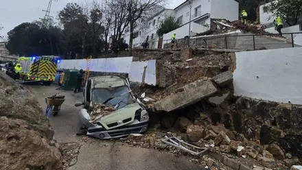 Imagen de varios bomberos actuando en una calle estrecha de Ubrique, junto a una vivienda parcialmente derrumbada tras el desprendimiento de una gran roca de la montaña. Los efectivos trabajan entre escombros, con un vehículo dañado en la calzada, mientras aseguran la zona durante el temporal.