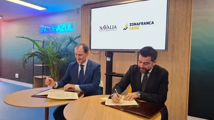 Fran González y Javier Arnau durante la firma del acuerdo en un entorno portuario, con elementos marítimos de fondo y logotipos de Zona Franca y Navalia visibles.