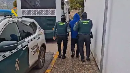 Un detenido con chubasquero azul esposado y conducido por dos agentes de la Guardia Civil.