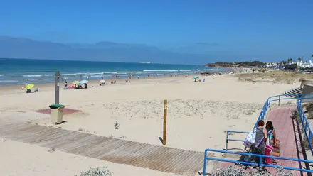 Playa de Chiclana con un suelo parcial de madera, luchas y mucha arena dorada, al fondo el mar en azul.