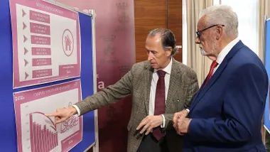Román y Maeztu, de chaqueta ambos, miran un panel explicativo en color morado y beige.