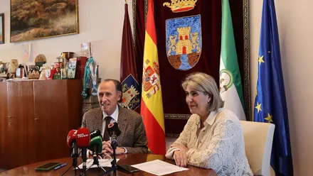 Román y Rico sentados en una mesa con las banderas española y andaluza tras ellos y el escudo de Chiclana.