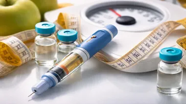 Jeringuilla cargada con un fármaco de color caramelo, detrás una báscula y un metro.