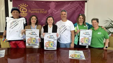 Miembros de la organización y la alcaldía posan con el cartel y el fondo en burdeos con letras doradas de Puerto Real.