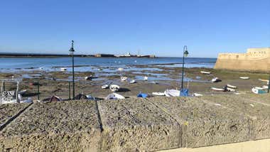 Bajamar en La Caleta con barquitas en las rocas y el Castillo de San Sebastián al fondo bajo un cielo azul en una imagen tomada desde la balaustrada de piedra ostionera, que se ve en la foto