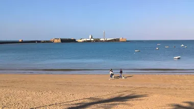dos personas pasean por con sus perros por la playa de la Caleta en un día de cielo azul y despejado. Foto: José Luis Porquicho Prada.