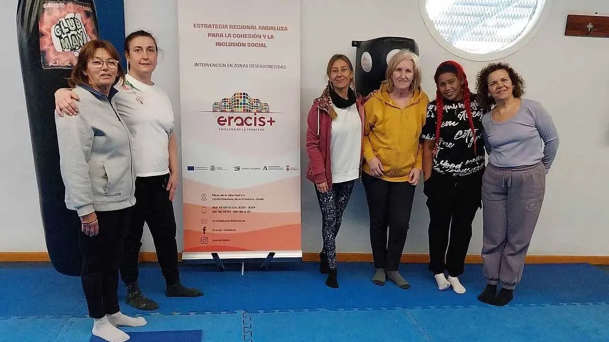 Mujeres del taller Eracis+ en la lona azul y posando junto al cartel.
