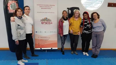 Mujeres del taller Eracis+ en la lona azul y posando junto al cartel.