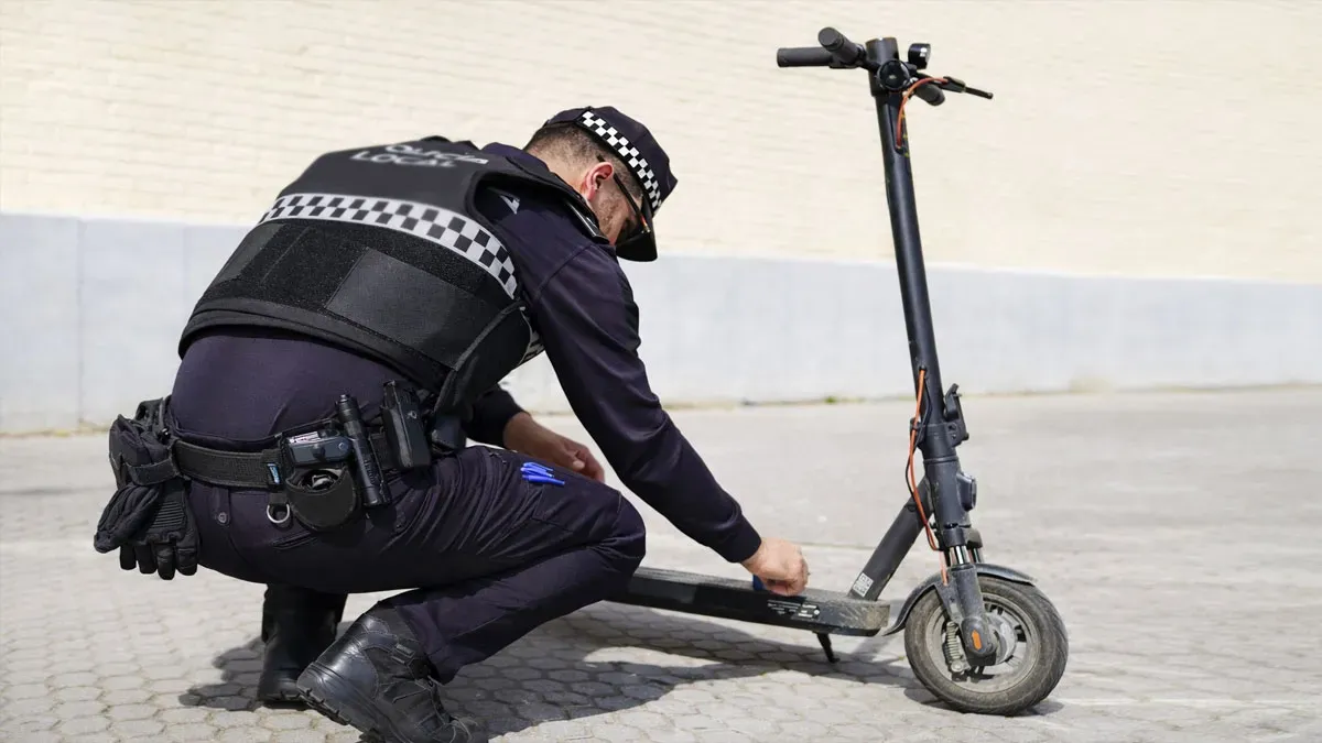 Policía local de azul agachado y verificando un patinete gris-negro.