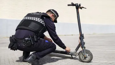 Policía local de azul agachado y verificando un patinete gris-negro.