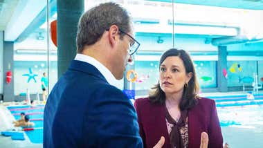 Patricia Cavada, con chaqueta color berenjena, habla cerca de la piscina con un hombre de chaqueta azul.