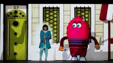 Actores en el escenario, uno de ellos disfrazado con una especie de pepino rosado.