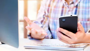 Persona con un teléfono en la mano delante de un ordenador.