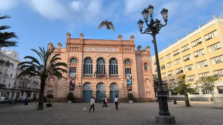 Varias personas camina por delante del Teatro Falla de Cádiz bajo un cielo mayormente despejado. Una paloma vuela en primer plano