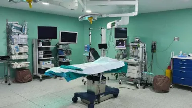 Mesa de operaciones e instrumental y aparatos de un quirófano. Destacan las paredes en un tono suave de verde.